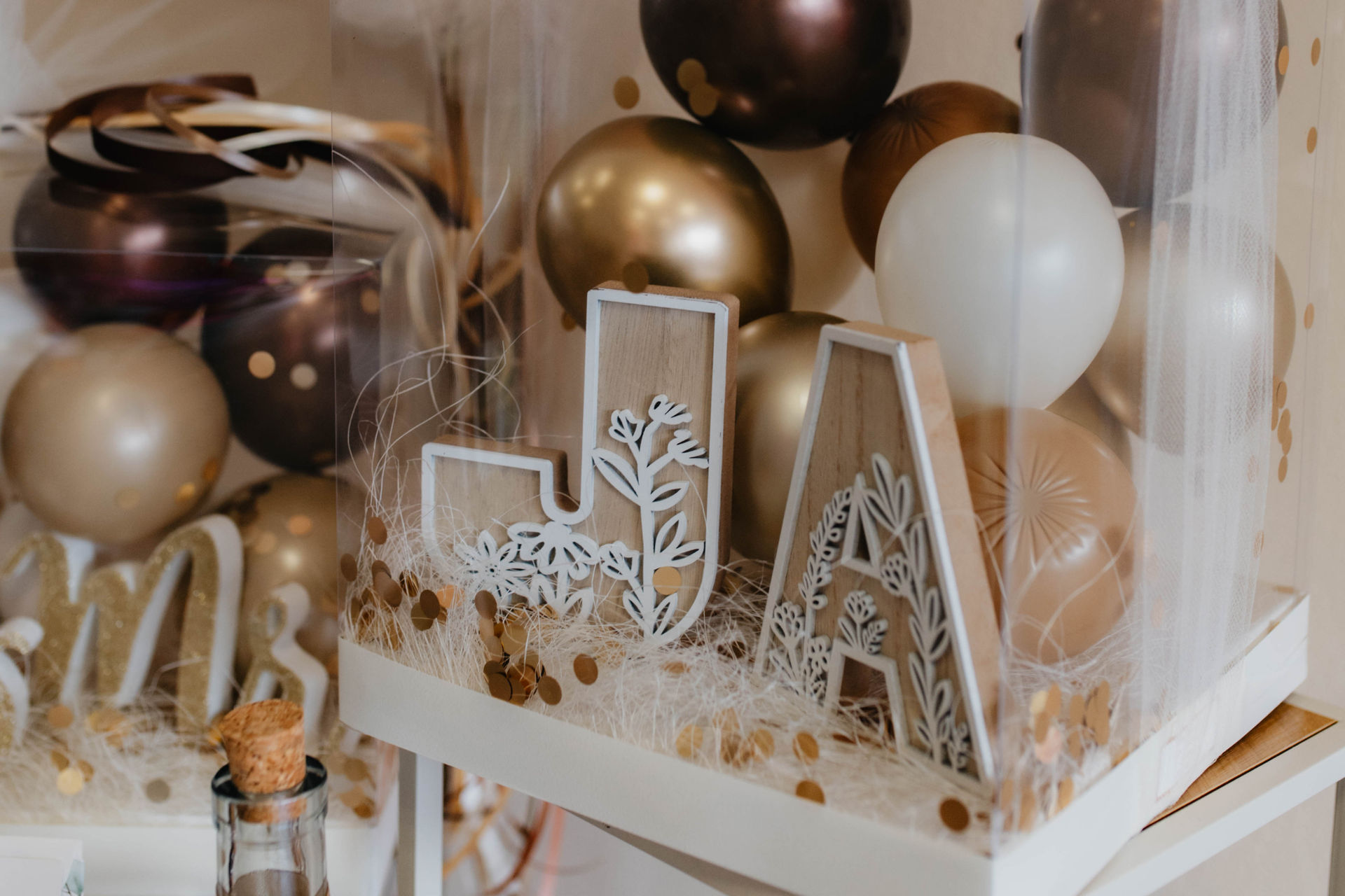 Holzbuchstaben mit weißen Mustern stehen vor goldenen und weißen Luftballons