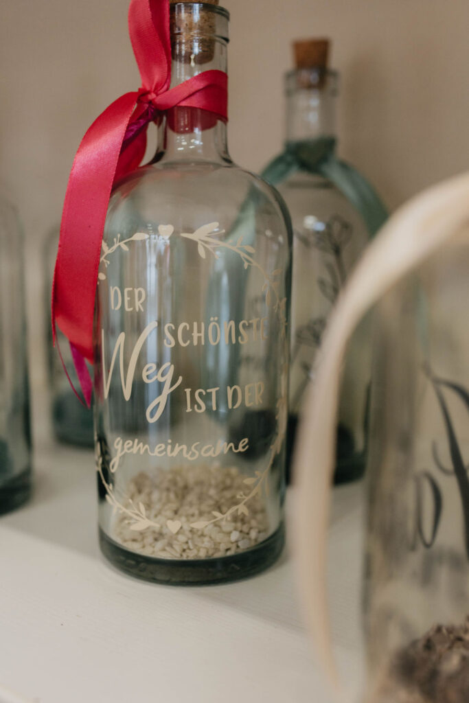 Eine Glasflasche mit rotem Band, Gravur und weißen Steinen im Inneren