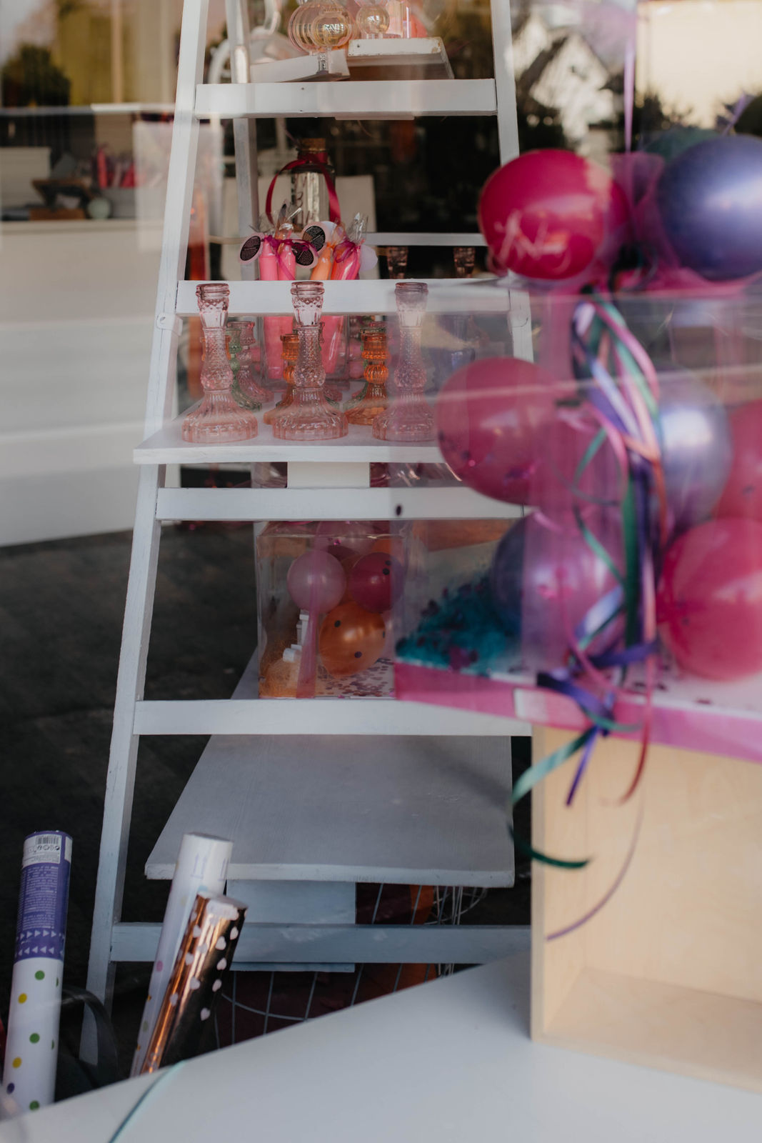 Mehrere bunte Ballons und Glasflaschen stehen auf einer weißen Leiter im Schaufenster