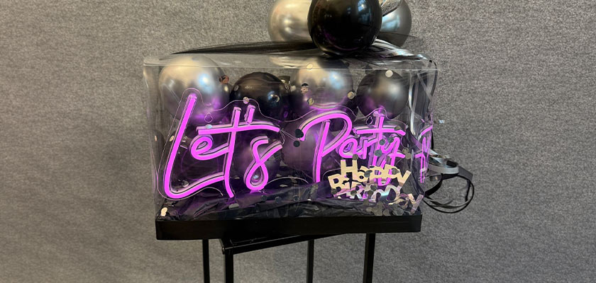 Ein leuchtendes pinkes Let’s Party Schild vor grauem Hintergrund mit schwarzen und silbernen Luftballons