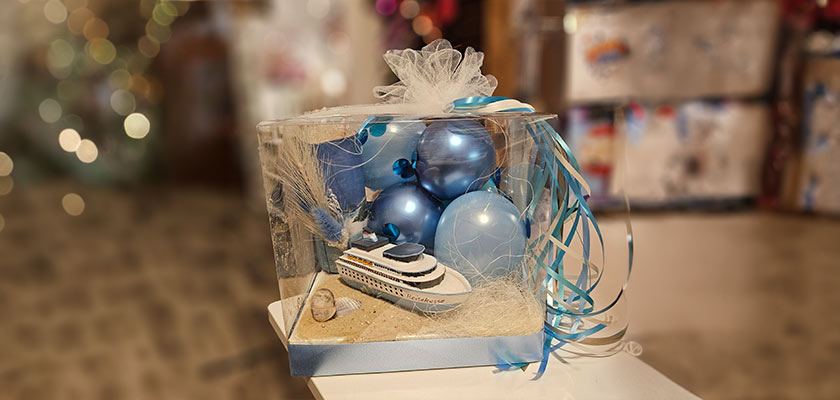 Ein Geschenk mit blauen Luftballons und einem Kreuzfahrtschiff als Modell in einer transparenten Schachtel